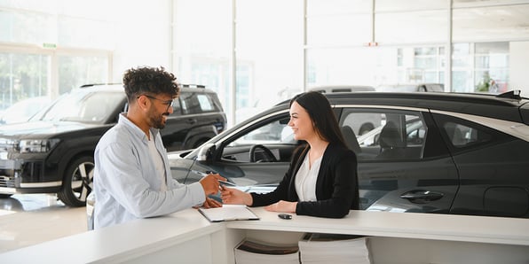A man and a woman stand at the counter of a large room. There is a clipboard with paperwork on the counter. They exchange a pen and there are cars in the background.
