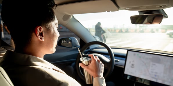 A man sits behind the drivers wheel in a vehicle and holds a tumbler cup in one hand